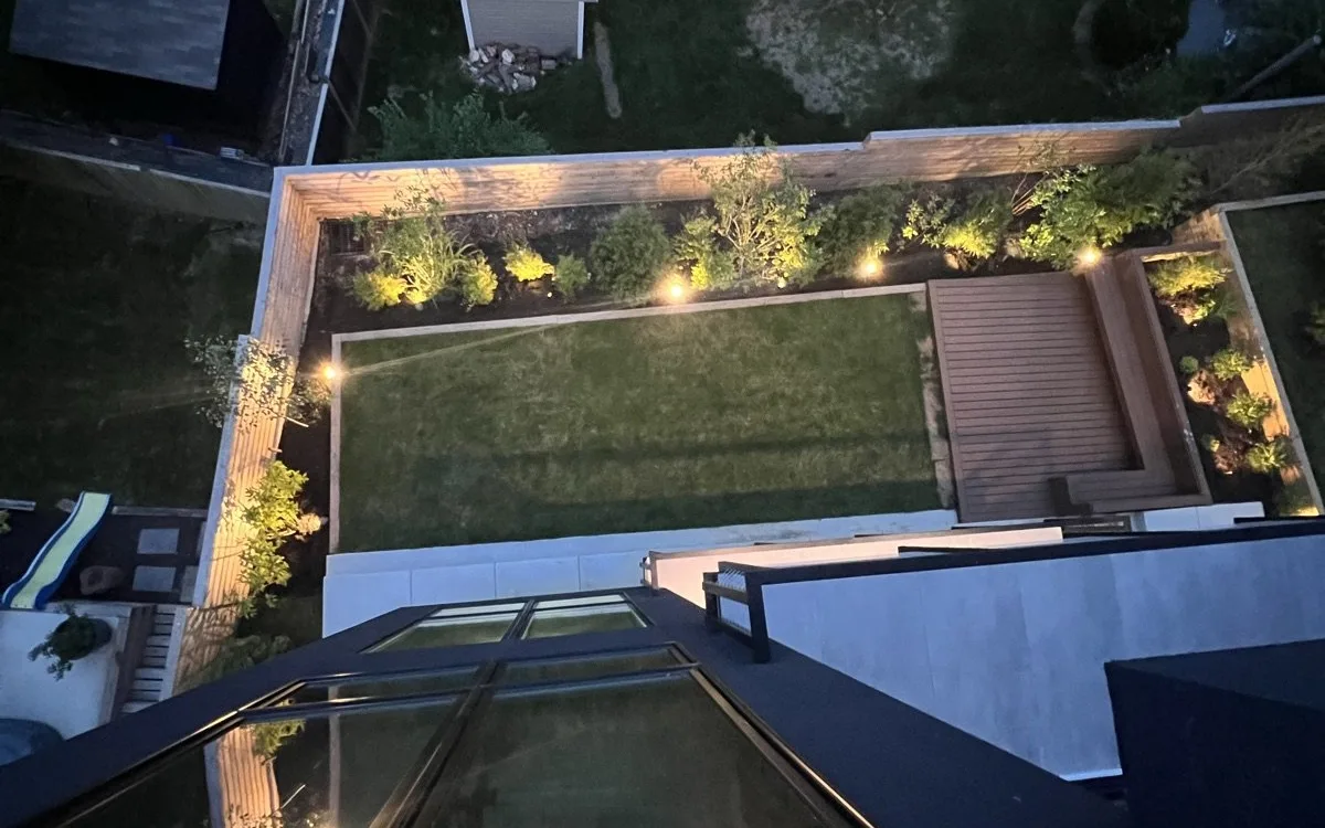 Aerial evening view of a renovated urban backyard with composite deck, raised cedar planting beds with landscape lighting, turf lawn, and large-format concrete paver patio steps