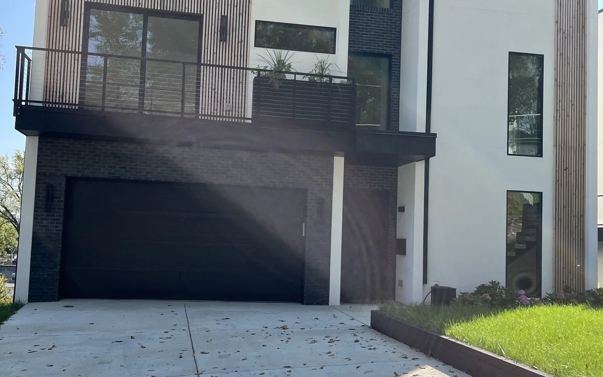 Contemporary home exterior renovation with smooth white stucco facade, dark vertical wood slat accent panels, large black aluminum casement windows, flat parapet roof, upper-level balcony with black steel railing, and matte black garage door
