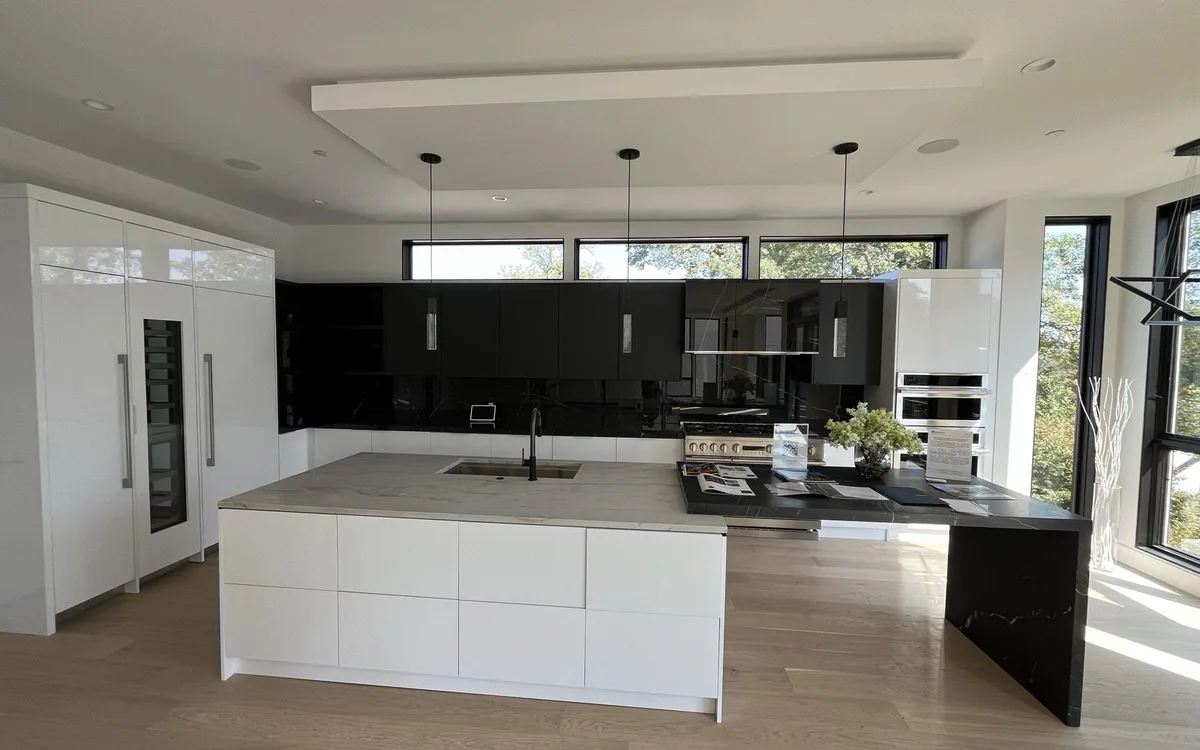 Contemporary kitchen with white flat-front handleless cabinets, large island with light stone countertop, dark stone waterfall extension, and light hardwood floors