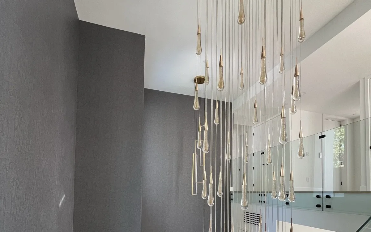 Cascading multi-pendant chandelier with dozens of teardrop glass and brass pendants hanging through a two-story foyer void, with glass panel stair railing visible and gray accent wall behind