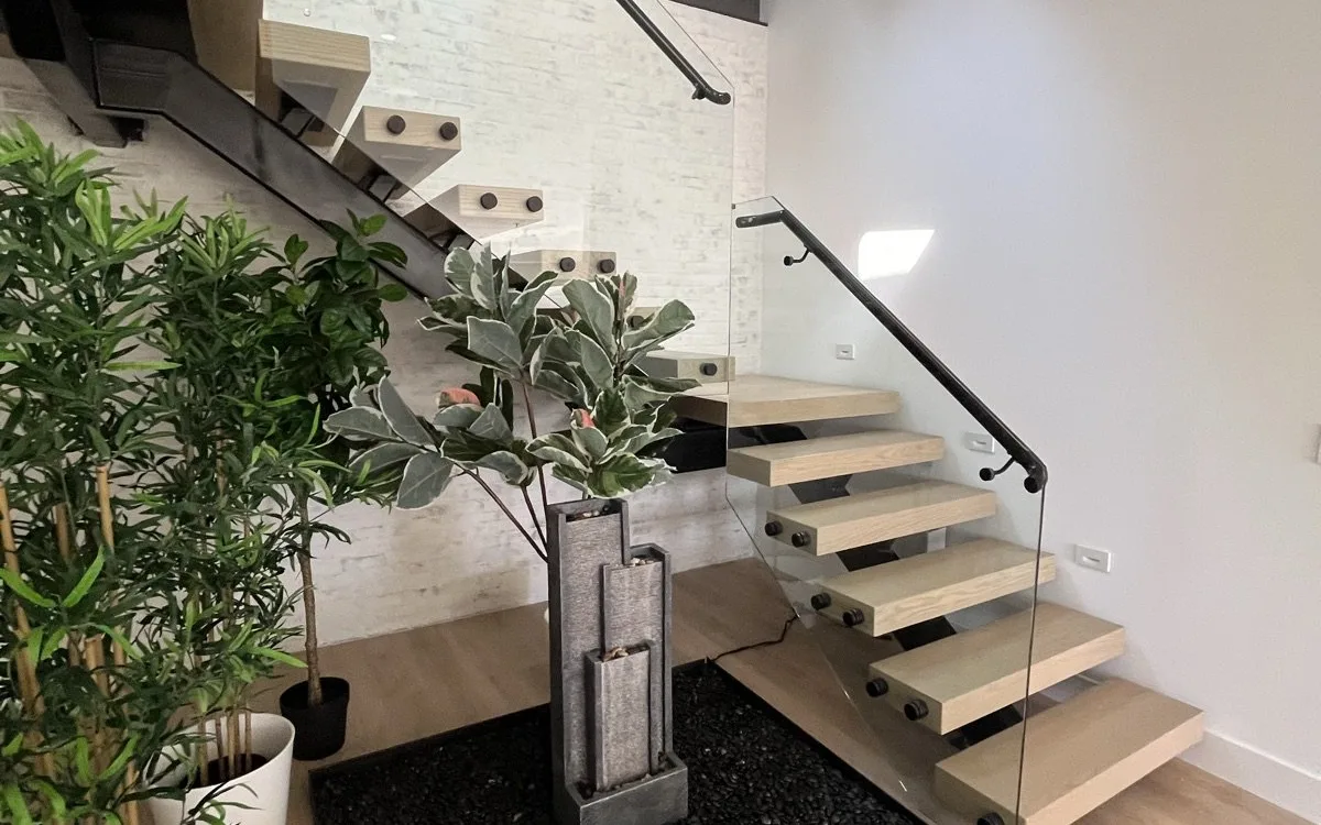 Floating open-riser staircase with glass railings above a designed under-stair indoor garden with black river rock, indoor water fountain, and potted plants